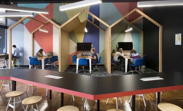 A vibrant contemporary collaborative workspace with multiple timber-framed, house-shaped booth structures arranged along a shared black workbench. Several people are working at desks with blue office chairs, computers, and stools. The space features bold geometric wall panels in red, blue, olive, and pale tones, exposed dark ceiling elements, linear pendant lighting, and a polished timber floor.