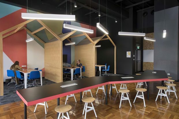 A contemporary commercial communal dining and work area, likely in a café, workplace, or educational facility. The space features a long black communal table with a vivid red edge, numerous white-legged timber stools, blue chairs, small tables, timber booth partitions, and distinctive timber-framed pitched canopy structures. Suspended linear pendant lights hang from a dark exposed ceiling, while bold red, blue, green, and black wall surfaces create a colourful, energetic interior.