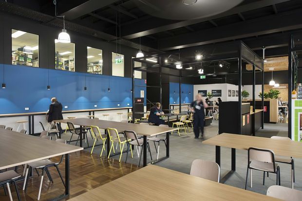 A large contemporary commercial interior, likely a café, food hall, or communal dining venue, with rows of timber-look tables and mixed chairs. The space features a bold blue partition wall, black exposed ceiling structure, suspended pendant lights, polished concrete flooring, glazed internal partitions, and a service or counter area toward the rear. The open layout and abundant seating create a bright, functional communal atmosphere.