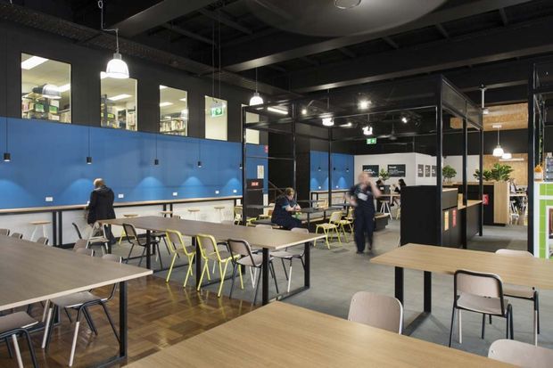 A large contemporary commercial interior resembling a school, workplace, or community dining and collaboration area. The open-plan space contains rows of timber-look tables, mixed chairs, pendant lights, blue partition walls, black exposed ceiling services, glazed meeting or classroom rooms, and visible occupants. The fit-out combines practical communal seating with a bold blue and black colour scheme.