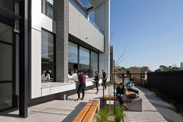 A contemporary multi-storey building courtyard or terrace with dark brickwork, white and grey panel cladding, extensive glazing, and a covered outdoor walkway. People are gathered around a built-in outdoor seating area beside planter beds and timber benches, with a black metal fence and distant urban buildings beyond.