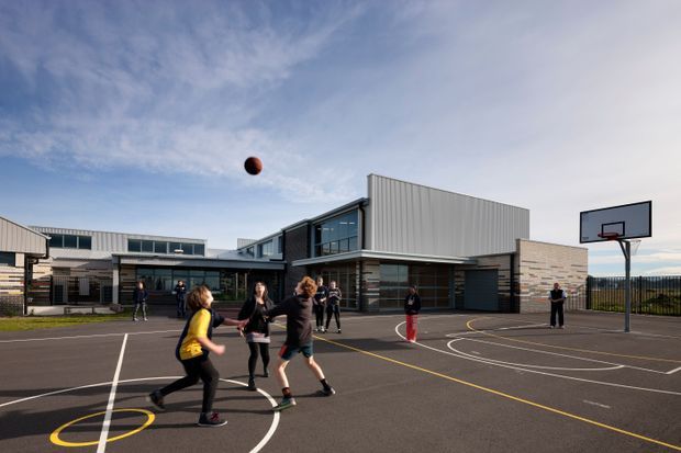 Exterior view of a contemporary school or community sports facility with a large outdoor basketball court in the foreground. The building has crisp geometric forms, extensive glazing, pale grey corrugated metal cladding, and light stone or brick walls. Basketball hoops, court markings, and people playing are visible under a partly cloudy sky.