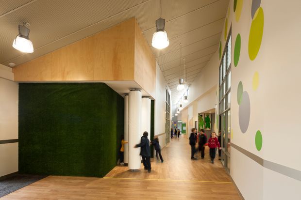 A bright, contemporary institutional hallway with warm timber flooring, a suspended timber-clad ceiling volume, white cylindrical columns, exposed glazing, and colourful circular wall graphics. People are present in the corridor, which appears to be a school or other public interior. A dark green textured feature wall and open circulation space define the foreground.