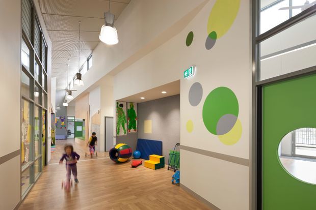 A bright, double-height institutional hallway, likely in a school or childcare facility, with warm timber flooring, large windows, glazed classroom fronts, pendant lights, colourful geometric wall graphics, and children's play equipment. Green doors and visible children reinforce the educational or childcare setting.