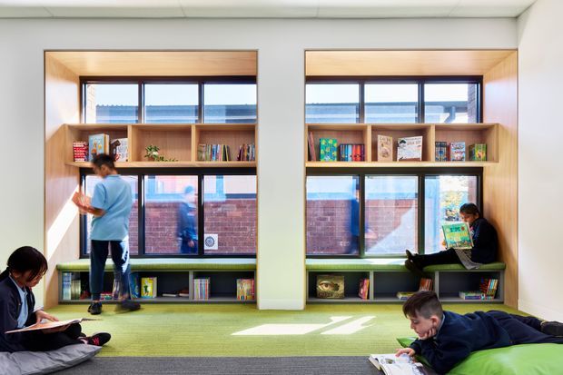Bright contemporary reading room or school library with two large window bays framed by built-in light-wood shelving. The shelves contain books and small educational items, with green upholstered or carpeted window seating below. Several children are reading or browsing, and sunlight creates a calm, welcoming learning environment. A brick building is visible outside through the glazing.