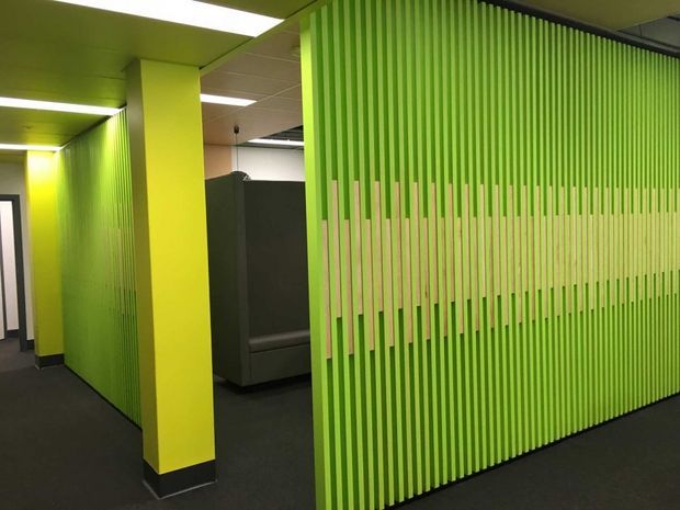 Interior view of a contemporary commercial workspace or office with vivid lime-green painted walls and columns. A prominent full-height screen of closely spaced vertical slats creates a semi-transparent partition, with a dark upholstered booth or seating area visible behind it. Dark carpet, recessed ceiling panels, and fluorescent-style ceiling lighting complete the utilitarian office fit-out.