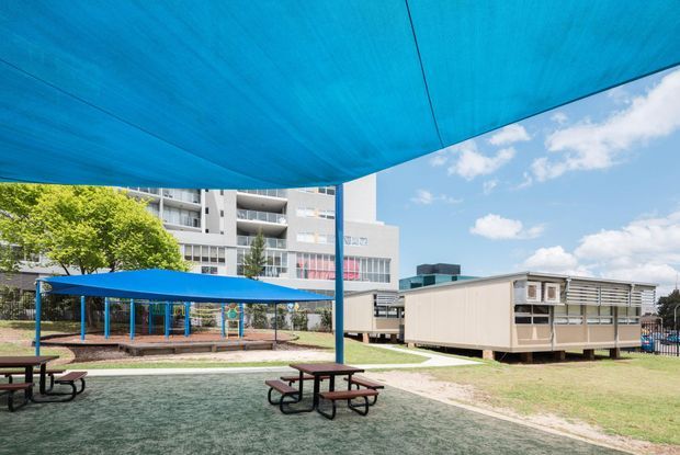 A covered outdoor recreation area with a large blue shade canopy, picnic tables, and a sandy landscaped surface. Additional blue shade structures, a portable classroom-style building, and a multi-storey institutional or apartment building are visible in the background under a bright sky.