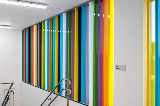 Interior view of a commercial stairway or landing with stainless-steel handrails, fluorescent ceiling lights, and a striking floor-to-ceiling feature wall composed of narrow vertical panels in vivid blue, green, purple, orange, yellow, and other bright colours. A small level sign is mounted on the white wall.