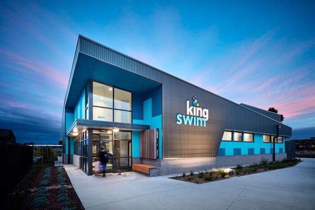 Exterior architectural photograph of a contemporary commercial swimming facility at dusk. The building has a large angular projecting canopy, tall glazed entrance, blue accent walls, ribbed dark-grey metal cladding, and prominent King Swim signage. A paved forecourt, landscaped planting, and a fenced side area are visible.