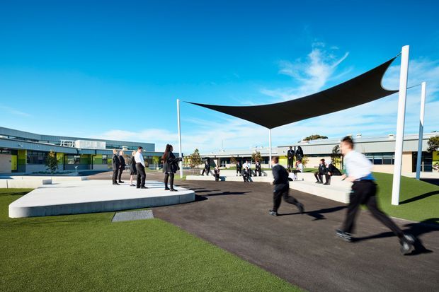A bright, contemporary landscaped courtyard at a low-rise institutional or commercial complex. The open paved space contains sculptural white concrete seating platforms, a large black tensile shade canopy supported by slender poles, synthetic-looking green lawn, and people walking or gathering. Long glazed and metal-clad buildings frame the courtyard in the background.