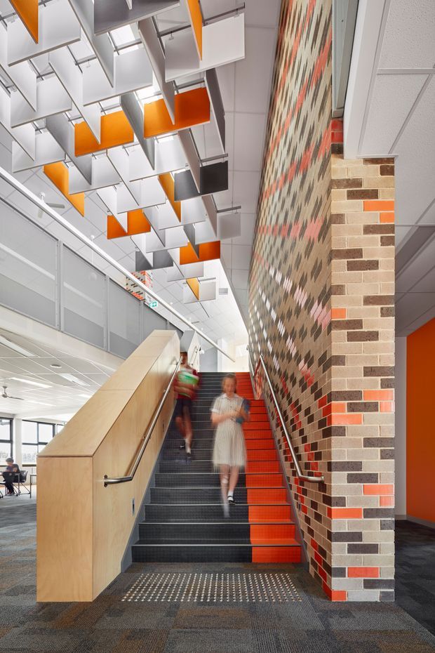 A contemporary commercial interior showing a stairway in an open multi-level circulation space. The stairs have dark carpeted treads with vivid orange risers, pale timber balustrade cladding, and stainless-steel handrails. A tall feature wall beside the stairs is constructed from varied brown, grey, cream, and orange bricks in a patterned composition. Above, suspended white, grey, and orange acoustic ceiling panels create a geometric installation beneath a tiled ceiling. Office glazing, carpet flooring, and blurred people reinforce the institutional or workplace setting.
