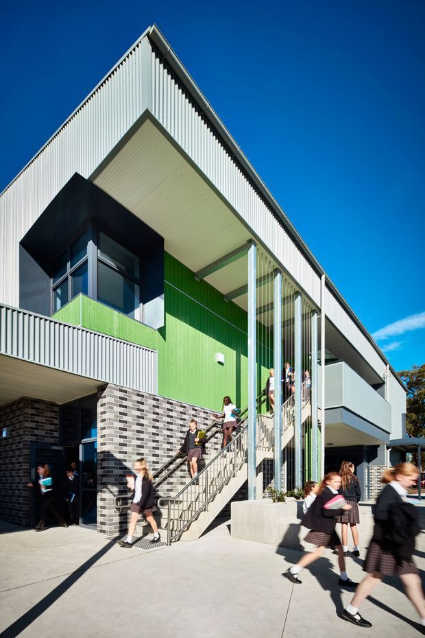 Exterior architectural photograph of a contemporary multi-storey school or institutional building. The facade combines white vertically ribbed metal cladding, dark grey brickwork, bright green wall panels, large glazed openings, projecting balconies, and a broad covered entrance canopy. An external metal stair with slim vertical screening leads to upper levels, with students moving through the foreground. Clear blue sky and strong sunlight emphasize the building's angular roofline and modern educational character.