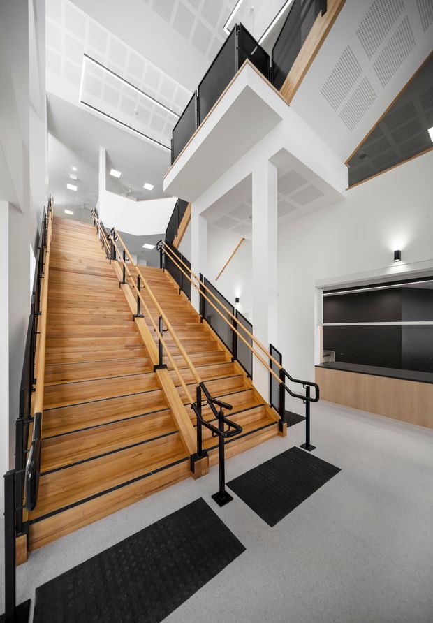 A bright, contemporary multi-level stairway and landing within a commercial or institutional interior. The broad timber staircase has black metal handrails and balustrades, with white walls, exposed black structural elements, perforated acoustic ceiling panels, and a light grey floor. A dark built-in service or reception counter is visible to the right beneath an upper-level walkway.