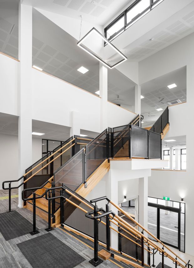 A bright, contemporary multi-level interior stairway and landing in a spacious institutional or commercial building. Multiple flights of stairs with black metal mesh balustrades, timber handrails and timber stair treads connect open mezzanine-like levels. White structural columns and walls, perforated acoustic ceiling panels, high-level glazing, dark carpeted landings and geometric suspended LED lighting create a clean, modern architectural interior.