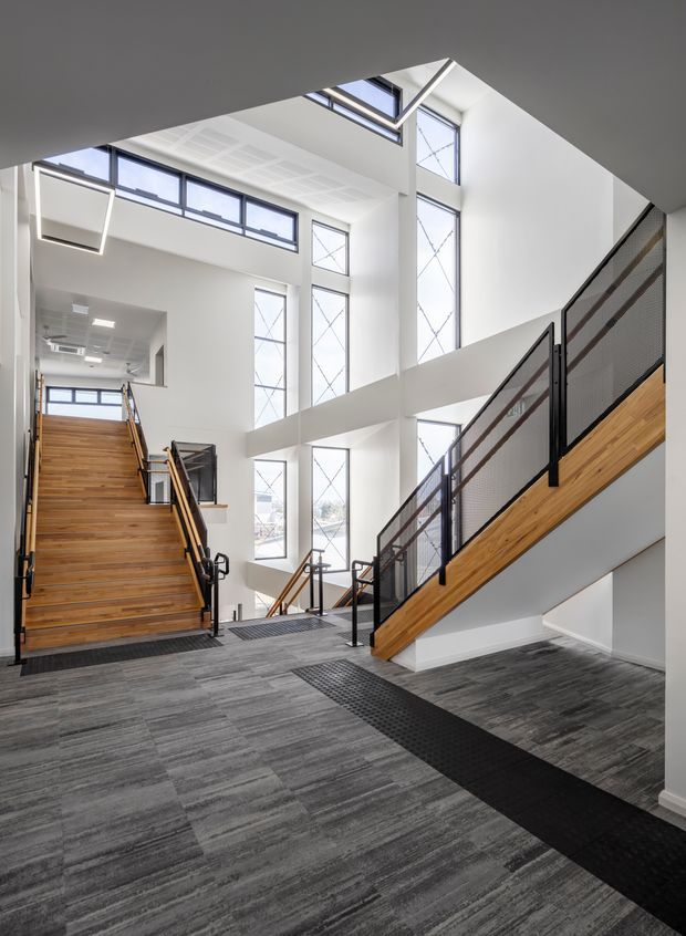 A bright, multi-level interior stairway and landing with a dramatic double-height void. The space features broad white walls, extensive tall glazing and clerestory windows, grey carpet tiles, and multiple timber stair flights with black metal and mesh balustrades. The composition emphasizes circulation, open volume, and abundant natural light.