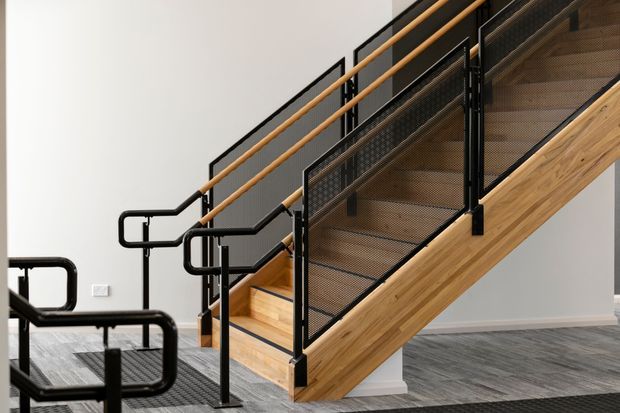 Interior view of a contemporary stairway with warm timber stair treads and stringer, black metal mesh balustrades, and matching black handrails. The stairs rise through a bright neutral space, with a grey textured floor and an additional black metal railing structure in the foreground.