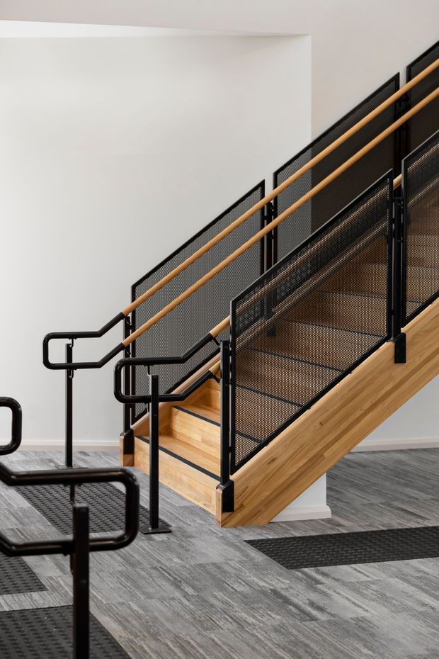 Interior view of a contemporary stairway with warm timber treads and side stringer, black metal handrails, and black perforated mesh balustrades. The stair connects to an upper landing, set against plain white walls and a grey patterned floor covering, with black entry mats at the base.