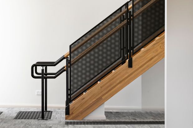 Interior view of a modern stairway with a warm timber stair stringer and treads, black metal handrails, and perforated black metal balustrade panels. The staircase rises diagonally against pale walls, with grey patterned flooring and a restrained contemporary finish.