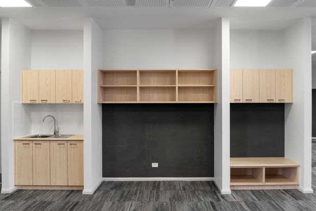 A clean, contemporary built-in office or utility workspace with pale timber cabinetry, open shelving, dark charcoal wall panels, and a grey patterned carpet floor. The left side includes a compact sink and benchtop, while the right side has additional closed storage and low open shelving. White structural columns and a white acoustic ceiling complete the functional commercial-style fit-out.