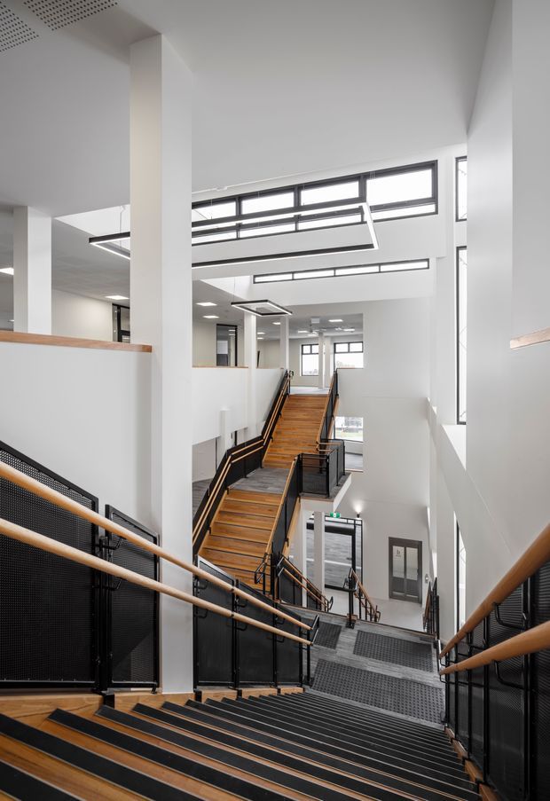 A bright, multi-level interior stairwell or circulation space with several flights of timber stair treads, black metal mesh balustrades, and timber handrails. White structural columns and walls surround the open stair void, with high clerestory-style glazing and linear suspended lighting creating an airy institutional or commercial atmosphere.
