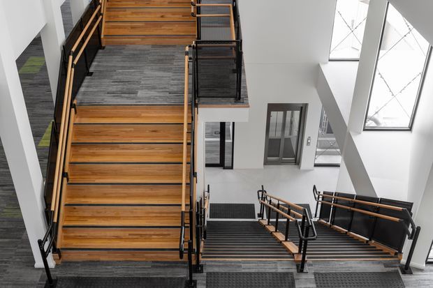 A contemporary multi-level stairway and landing viewed from above, featuring broad timber treads, dark metal handrails, black mesh balustrades, and grey flooring. White walls, tall glazing, and a bright double-height void create an open, industrial-inspired circulation space.