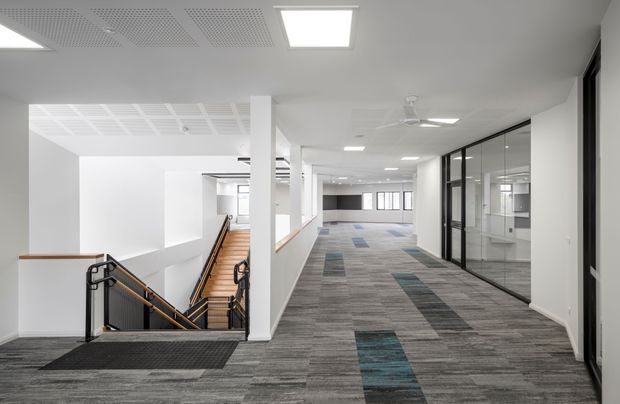 A bright, contemporary commercial interior showing a wide carpeted corridor beside a stairwell. The space has white walls and columns, acoustic ceiling panels with recessed LED lighting, ceiling fans, black-framed glazed office partitions, and grey patterned carpet with occasional blue-green inset strips. The stair has timber treads, dark metal balustrades, and a black entrance mat.