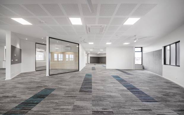A bright, open-plan commercial office interior with extensive grey modular carpet flooring, white walls, glazed meeting-room partitions, large windows, and a suspended perforated acoustic ceiling with recessed lighting. The space is mostly unfurnished, with a clean contemporary workplace fit-out and colourful linear carpet inlays.
