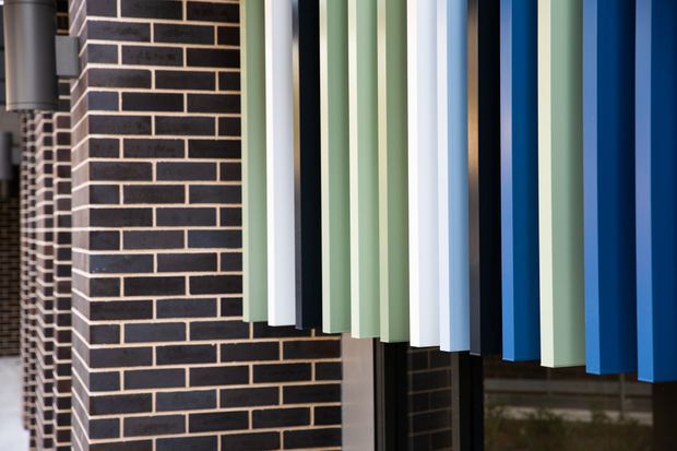 Close-up architectural detail of a dark brick facade with projecting vertical fins or louvres in alternating pale green, white, light blue, and deep blue finishes. Black-framed glazing and a dark metal cylindrical element are partially visible, creating a contemporary, graphic exterior composition.