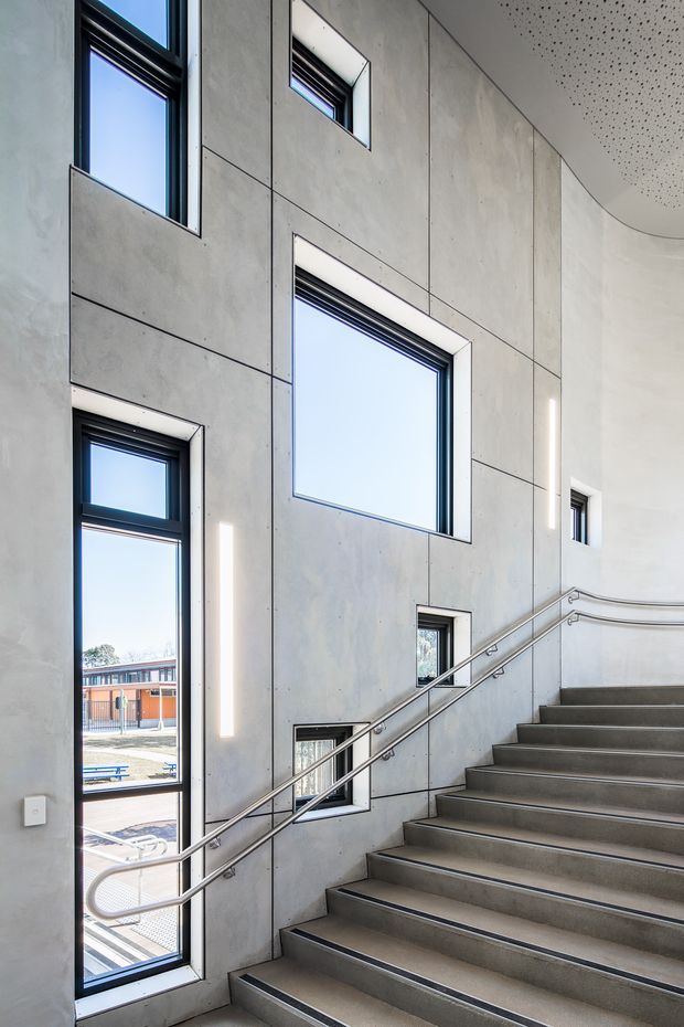 A bright contemporary stairwell with broad concrete stairs, stainless-steel handrails, and a tall wall of irregularly arranged black-framed windows. Large pale concrete wall panels with expressed joints rise to a curved perforated ceiling, creating a clean institutional or commercial interior with strong natural light.