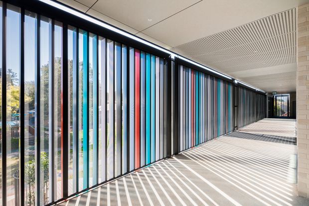 A long, bright covered architectural corridor with floor-to-ceiling glazed openings and closely spaced vertical coloured fins or panels along one side. Strong striped sunlight and shadows fall across the pale floor, while the ceiling features linear acoustic slats and recessed lighting. A light masonry wall frames the right side, and landscaped surroundings are visible through the glazing.