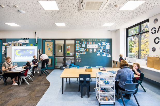 Bright contemporary primary-school classroom with students and teachers working at grouped tables. The room features teal and dark grey built-in cabinetry, an interactive display, educational wall boards, large windows, carpeted and vinyl flooring zones, acoustic ceiling tiles, and recessed LED lighting.