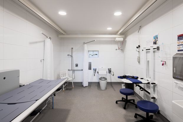 Bright, clean clinical examination and treatment room with white tiled walls, a large padded treatment table, medical equipment, privacy curtains, stools, wash basin, and recessed ceiling lighting.