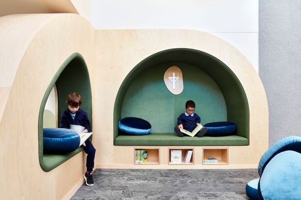 A playful, sculptural reading and retreat area designed for children, featuring curved pale timber wall joinery with deep green upholstered alcoves, built-in bench seating, blue cushions, and integrated open book shelving. Two children are reading in separate niches. The space has a soft grey carpet, rounded architectural forms, and a colourful, welcoming educational or library-like atmosphere.