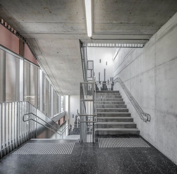 A raw concrete interior stairwell with intersecting flights of stairs, metal handrails, perforated metal balustrades, and an industrial linear light. The space is bright and highly geometric, with exposed concrete walls and ceiling, dark speckled flooring, and translucent patterned glazing or screens along the left side.