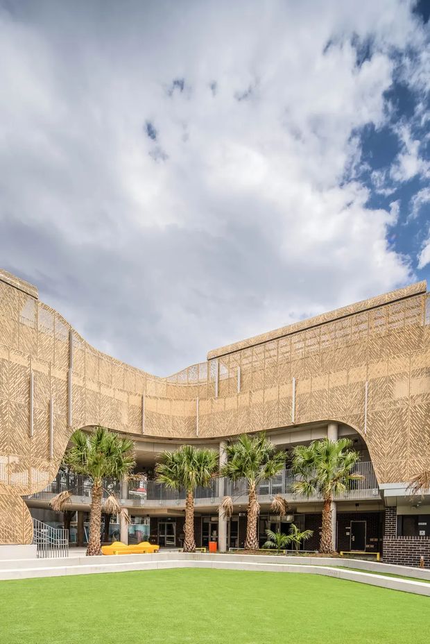 Exterior architectural photograph of a contemporary multi-storey building enclosing a landscaped courtyard. The facade is wrapped in a distinctive perforated or patterned tan screen, with curved balcony edges, exposed circulation elements, dark brickwork at ground level, palm trees, and a bright green lawn. A partly cloudy sky is visible above.
