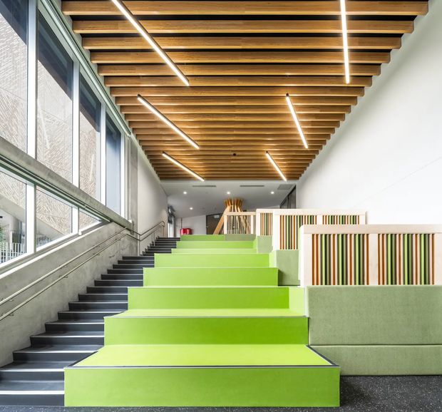 A bright contemporary interior circulation and gathering space with broad lime-green stepped seating beside a dark stairway. The space features full-height glazed windows with patterned screening, a warm timber slatted ceiling with integrated linear lighting, pale walls, green upholstered bench seating, and decorative vertical timber-and-colour screening around an upper landing.