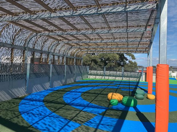 A covered outdoor recreational court or play area with a curved perforated metal canopy, steel fencing, padded orange posts, and blue and green sports surfacing. Several soft play or sports cushions lie on the court. Strong patterned shadows from the canopy create a graphic, high-contrast appearance, with trees and sky visible beyond the enclosure.
