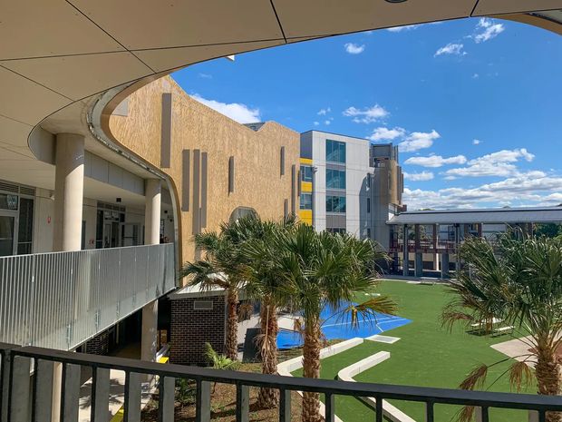 View from an elevated covered walkway or balcony across a contemporary multi-storey complex, with curved roof canopies, textured tan masonry walls, glazed vertical sections, metal railings, palm landscaping, and a landscaped courtyard featuring bright blue recreational surfaces and artificial turf. Blue sky and neighboring building wings are visible in the background.