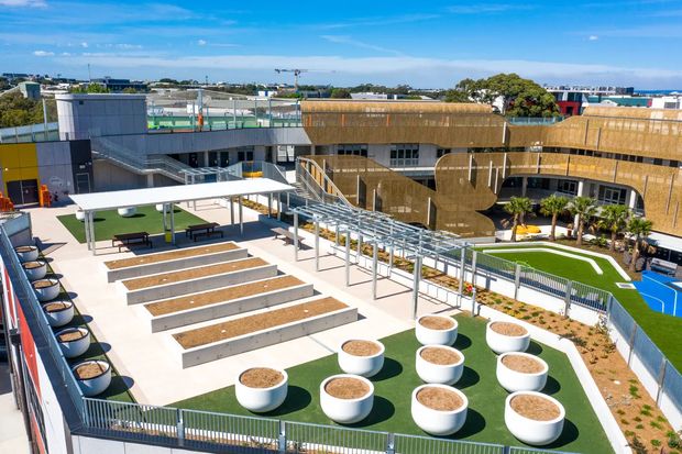 A bright elevated view of a contemporary educational or commercial campus courtyard, featuring tiered concrete seating steps, a white shade canopy, pergola-like walkway structure, artificial turf, large round white planters, landscaped planting, and a multi-wing building with curved facade elements. Sports courts and rooftop areas are visible in the background.