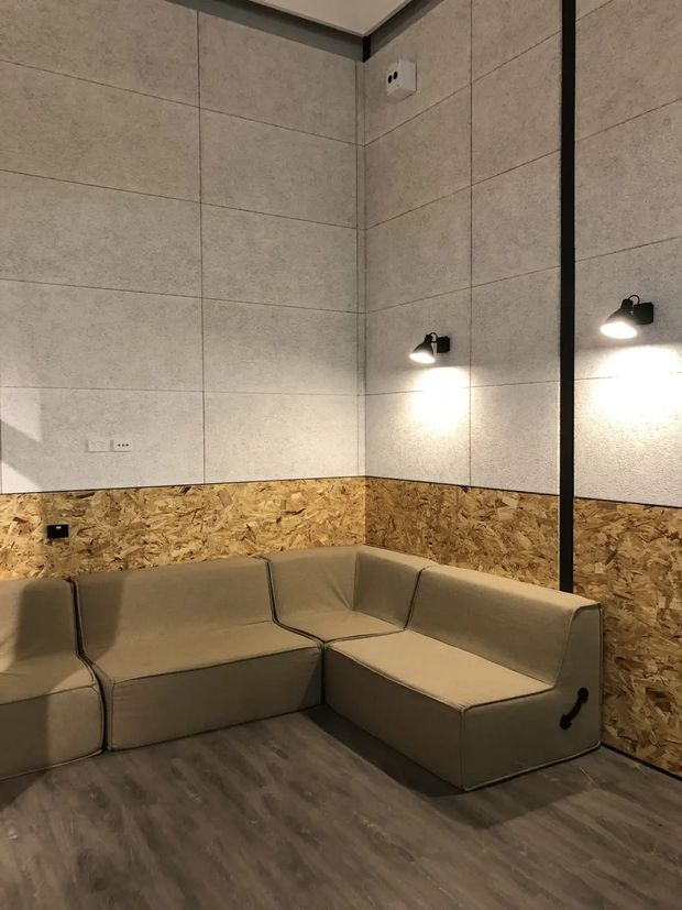 A compact lounge or media-room interior with a large L-shaped upholstered sectional sofa. The room features grey wood-look flooring, pale concrete-look wall panels, a band of exposed oriented strand board (OSB) wall lining, black-framed mirror or glazing, and simple black wall-mounted lights creating a warm, understated atmosphere.