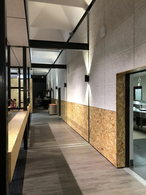 A long, modern commercial interior corridor with dark timber-look flooring, exposed OSB panels forming the lower wall sections, pale acoustic or fibre-cement wall panels above, and black steel-framed glazed partitions and doors. Linear black beams and framing create a strong industrial grid overhead, while wall-mounted up-and-down lights illuminate the passage. A kitchen or service area is visible through the doorway on the right, and work or dining areas appear farther down the corridor.