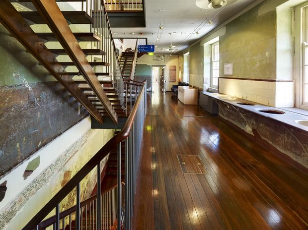 Interior view of a long, narrow multi-level space with exposed timber staircases, dark polished timber flooring, industrial-style metal balustrades, aged textured walls, and a tiled kitchenette or utility counter along the right side. Large windows provide natural light, while an open stair void and upper-level mezzanine create a strong vertical connection.