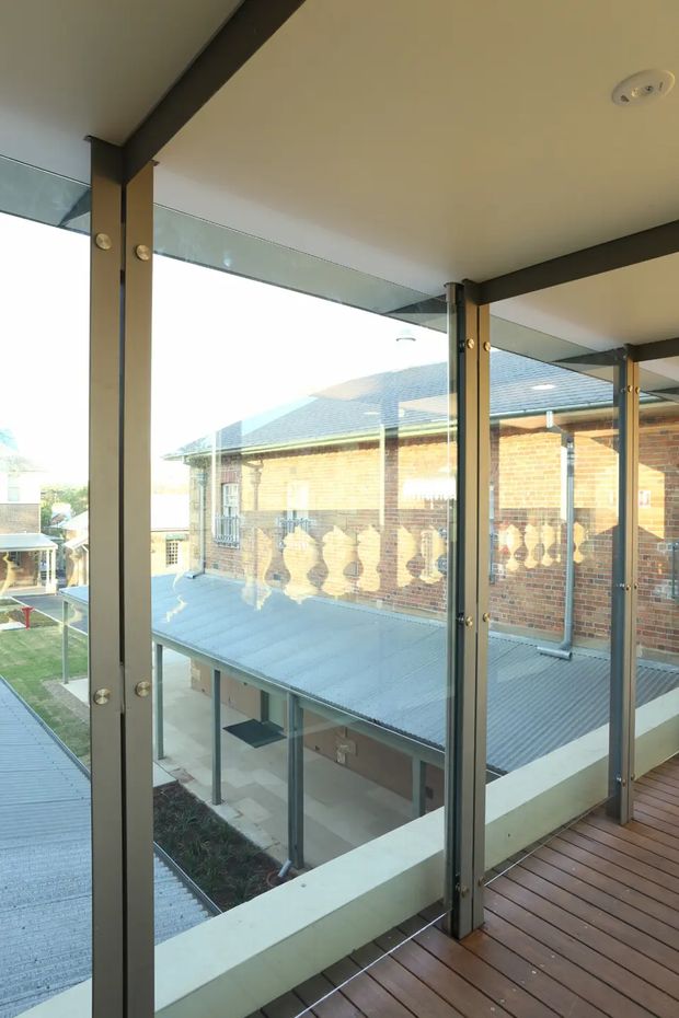 View from an elevated enclosed walkway or balcony through large clear glazing, overlooking a multi-level courtyard and adjacent brick building. The space has dark metal structural posts and framing, a timber plank floor, glass balustrade or walls, and a warm neutral ceiling with recessed lights. The surrounding building features brick masonry, corrugated metal roofing, and decorative balustrades.