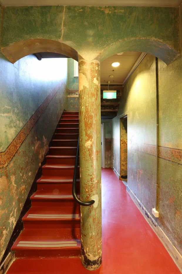 A narrow interior stairway and hallway with a strongly weathered, distressed finish. Red-painted stairs and flooring contrast with mottled green walls, a central cylindrical column, dark handrail, and decorative patterned wall trim. The corridor has a low ceiling with recessed lighting and visible peeling, layered paint, creating an intentionally aged and dramatic atmosphere.