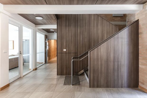 Modern interior hallway and stairway with a strong timber-lined stair enclosure, exposed brick wall, pale large-format tiled flooring, white structural beams, and glazed doors opening to adjacent rooms. The stair features sleek metal handrails and a partially concealed lower level, creating a refined contemporary circulation space.