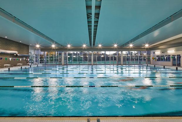 Large indoor swimming pool facility with multiple lap lanes, clear water, tiled pool surround, extensive floor-to-ceiling glazing, and a broad modern ceiling with a central linear skylight or clerestory. The space is brightly illuminated and appears to be a commercial or institutional aquatic centre.