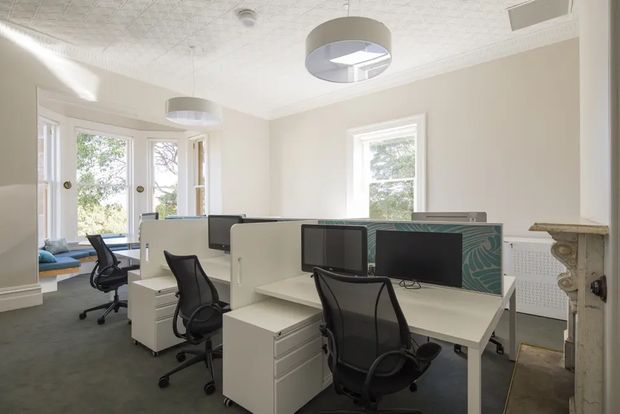 Bright, traditional office interior with multiple white workstation desks arranged in rows, black ergonomic mesh office chairs, computer monitors, and low white drawer units. The room has pale beige walls, dark carpet, tall white-trimmed windows and glazed doors, an ornate white pressed-tin ceiling, circular pendant lights, and a decorative carved fireplace surround. A small adjoining lounge area is visible through the opening at left.