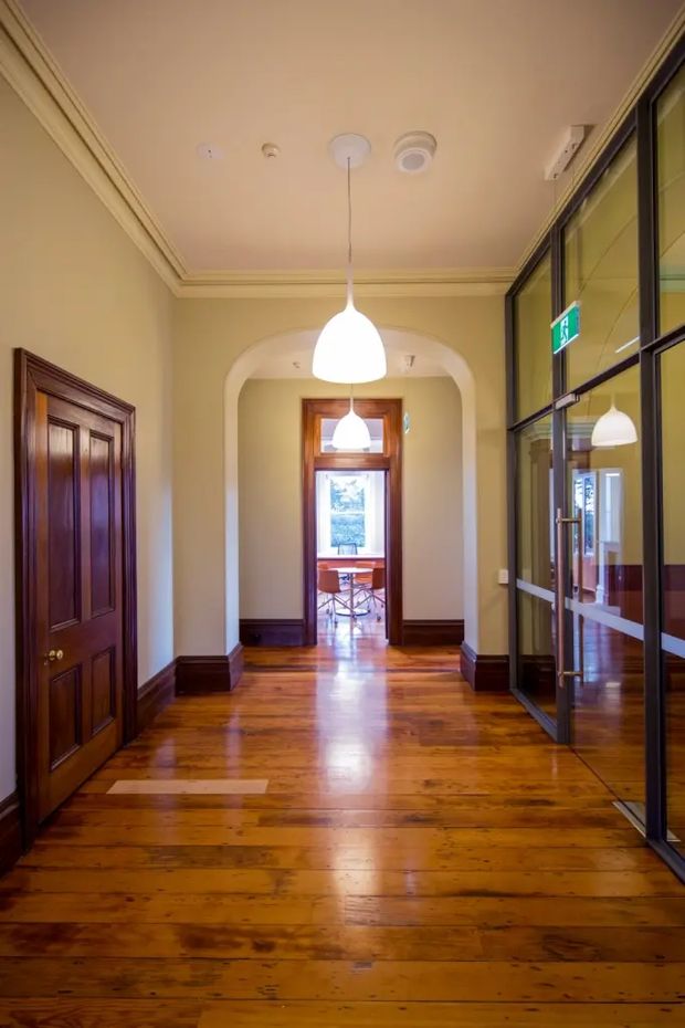 A polished heritage-style interior hallway with warm cream walls, ornate crown moulding, dark timber skirting, and glossy honey-toned timber floorboards. A richly stained timber panel door is on the left, while a black-framed glazed partition lines the right side, reflecting the corridor and pendant lighting. An arched opening leads toward a bright room with timber-framed glazing and seating beyond. Two white pendant lights hang along the circulation space.