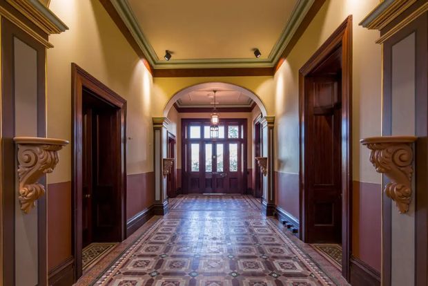 A grand, symmetrical residential hallway with richly detailed dark timber door surrounds, ornate carved corbels, wainscoting, layered cornices, and an arched opening leading to glazed entry doors. The patterned decorative tile floor and warm ochre, burgundy, and cream palette create a formal historic character.