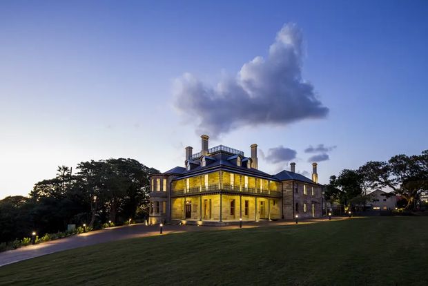 A grand historic two-storey mansion photographed at dusk, set within expansive landscaped grounds. The symmetrical dark masonry facade features tall illuminated windows, a projecting central entrance volume, classical proportions, multiple chimneys, and a decorative rooftop balustrade. Mature trees, a broad lawn, and a curved illuminated walkway surround the building.